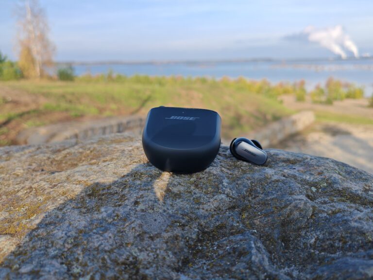 Die Bose QuietComfort Ultra Earbuds und ihr Ladeetui sitzen auf einem großen Felsen vor der malerischen Kulisse einer Graslandschaft und eines schimmernden Gewässers und bieten so die perfekte Umgebung, um ihre überragende Klangqualität inmitten der Ruhe der Natur zu testen.