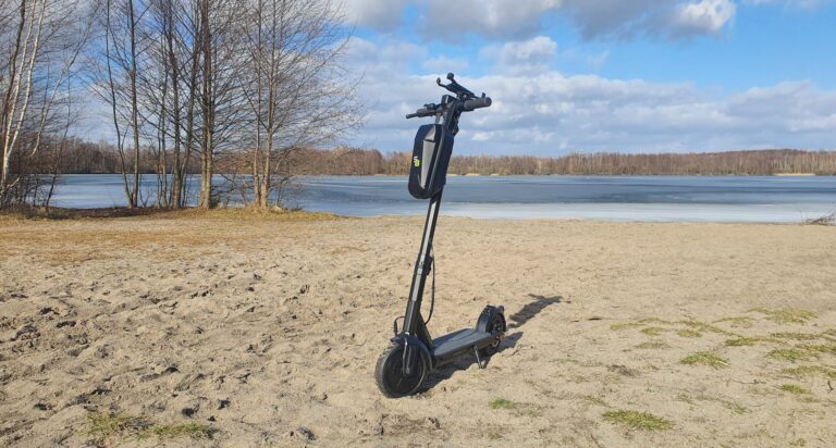 Elektroroller geparkt an einem Sandstrand neben einem teilweise zugefrorenen See, umgeben von kahlen Bäumen unter einem teilweise bewölkten Himmel.