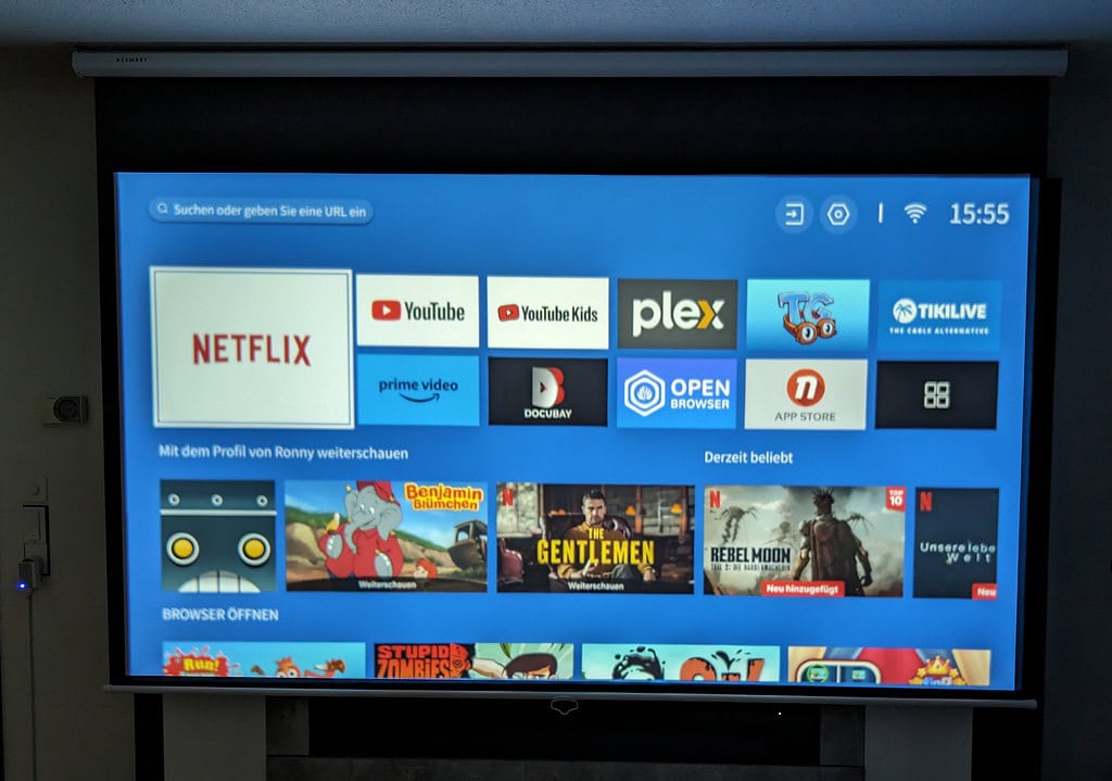 Auf einem Projektionsbildschirm wird vor blauem Hintergrund ein Home-Menü mit verschiedenen App-Symbolen angezeigt, darunter Netflix, YouTube und Prime Video. Die angezeigte Uhrzeit ist 15:55.