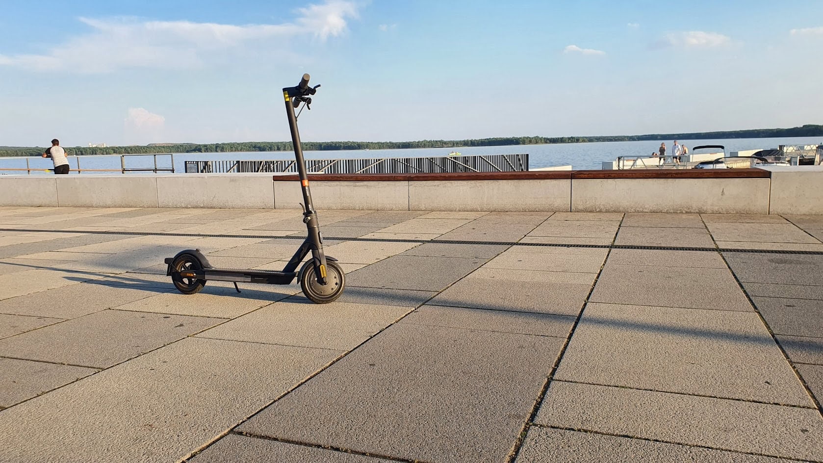 Ein Elektroroller ist auf einer gepflasterten Promenade am Wasser geparkt, im Hintergrund sind ein paar Boote zu sehen und der Himmel ist teilweise bewölkt.