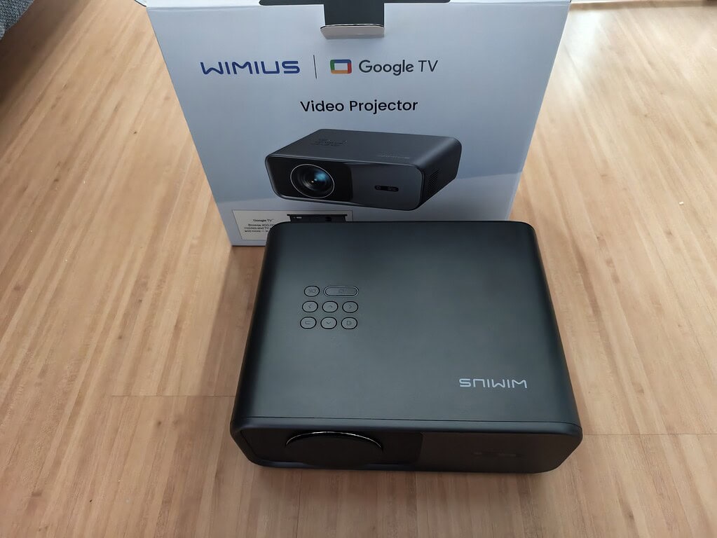 Ein WiMiUS G1 Test-Videoprojektor steht auf einem Holzboden vor seiner Box, auf der das Google TV-Logo und ein Bild des Projektors zu sehen sind.