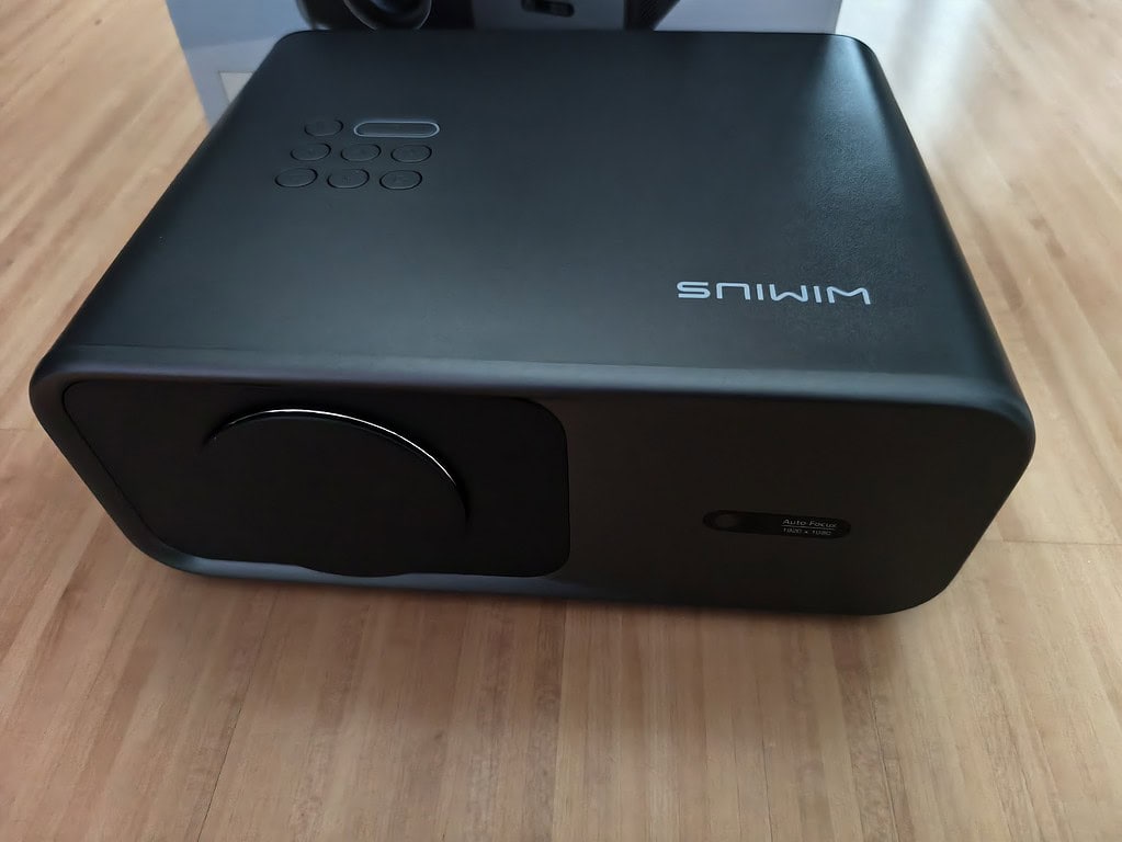 Ein schwarzer Projektor mit Objektivabdeckung steht auf einem Holzboden; das Wort "SMIWM" ist auf der Oberseite verkehrt herum aufgedruckt, was darauf hindeutet, dass es sich um ein WiMiUS G1 Testgerät handeln könnte.