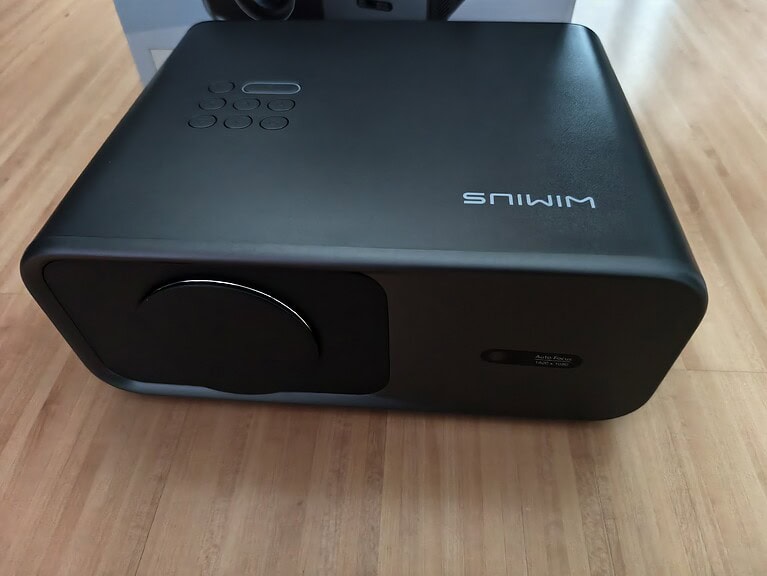 Ein schwarzer Projektor mit Objektivabdeckung steht auf einem Holzboden; das Wort "SMIWM" ist auf der Oberseite verkehrt herum aufgedruckt, was darauf hindeutet, dass es sich um ein WiMiUS G1 Testgerät handeln könnte.