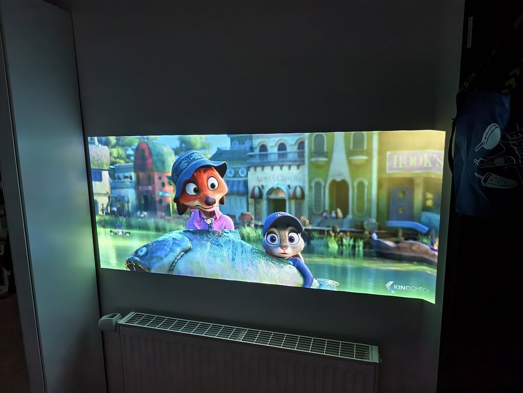 Ein WiMiUS G1 Test projiziert eine animierte Filmszene auf eine Wand über einem Heizkörper, in der zwei anthropomorphe Tiere vor bunten Gebäuden erscheinen.