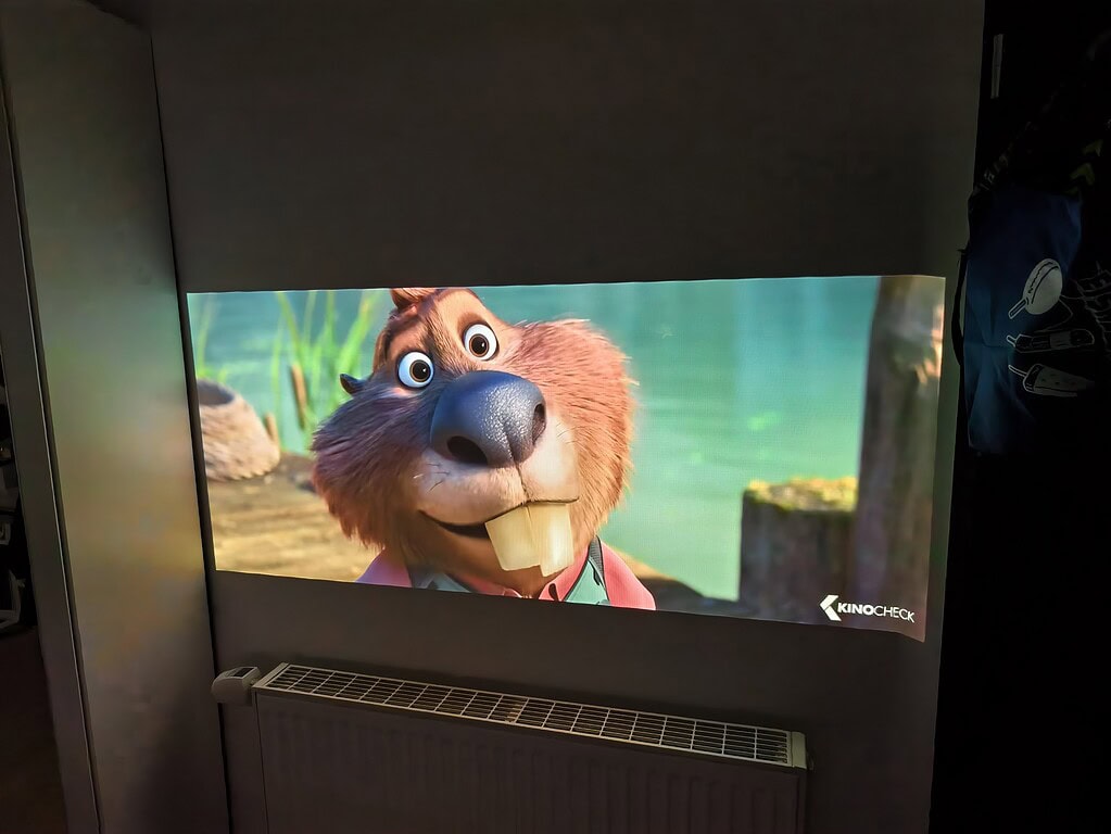 Eine animierte Biberfigur mit großen Zähnen erscheint während des WiMiUS G1-Tests auf einer Projektionsfläche über einem Heizkörper in einem schwach beleuchteten Raum. In der Ecke des Bildschirms ist ein Logo mit der Aufschrift "KINO CHECK" zu sehen.