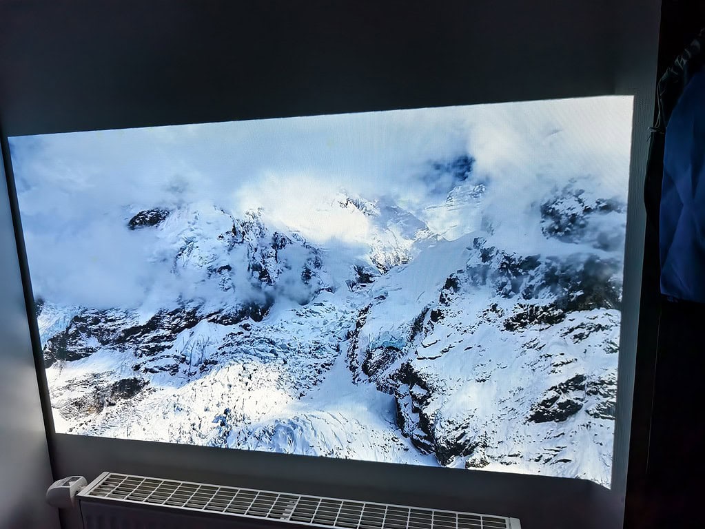 An der Wand über einem weißen Heizkörper ist ein großer Bildschirm angebracht, der eine Luftaufnahme von schneebedeckten Bergen und Wolken zeigt - perfekt für einen WiMiUS G1 Test, um atemberaubende Bilder zu erleben.